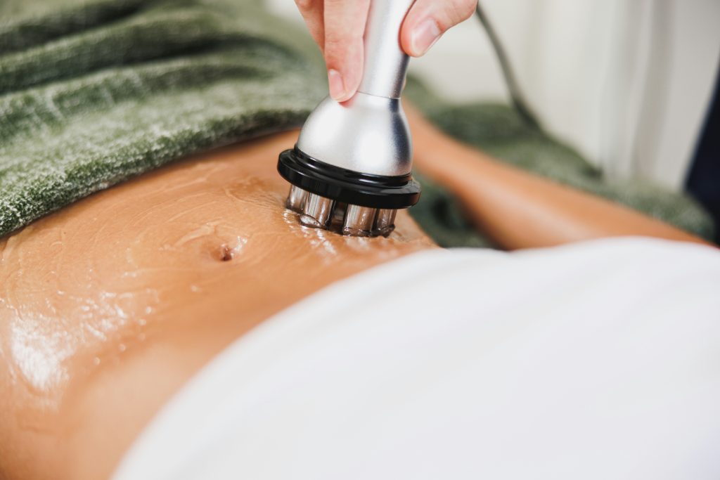 Hands guiding a metallic rolling massage device over a person’s oiled back for spa treatment.